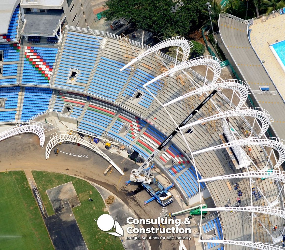 Vista aérea del Estadio Pascual Guerrero en construcción con asientos azules y rojos, vigas curvas blancas en proceso de instalación, vehículos de construcción y una gran piscina en las inmediaciones. El logotipo de Consulting & Construction aparece en la parte inferior.