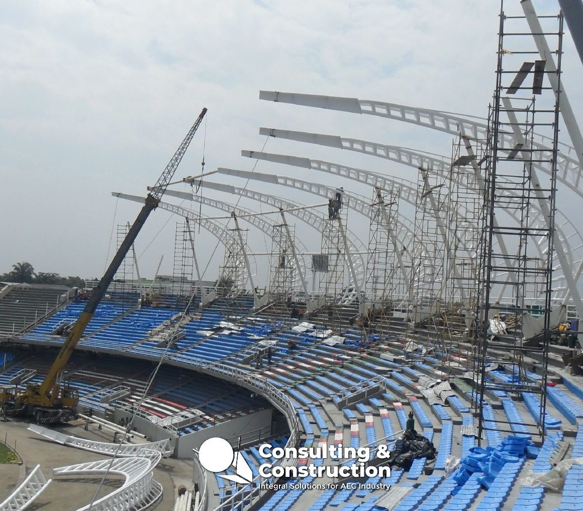 Trabajos de construcción en el Estadio Pascual Guerrero de Cali, con grúas y andamios montando la estructura curva del techo sobre las gradas azules y blancas. Se ven varios trabajadores en las obras del estadio.