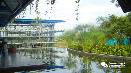 Un moderno edificio con estructura de acero vista, acentuada por Tintas Industriales, se alza tras un estanque reflectante y una exuberante vegetación. Por encima, plantas colgantes descienden del tejado. El logotipo de Consulting & Construction aparece en la esquina inferior derecha.