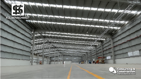 Vista interior de una gran nave industrial vacía con altos techos metálicos y una brillante iluminación cenital, que muestra el proyecto de Kolbenschmidt Ampliación Planta. Los logotipos de Kolbenschmidt y Consulting & Construction son visibles en las esquinas de la imagen.