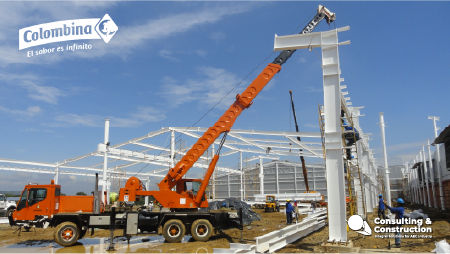 Una gran grúa naranja levanta una viga de acero blanca en las obras de Fase II para una estructura metálica. Se ven trabajadores y equipos. Los logotipos de Colombina del Cauca y Consulting & Construction aparecen en las esquinas superior izquierda e inferior derecha.
