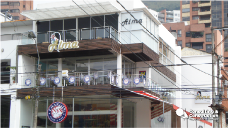 Un moderno edificio de dos plantas con grandes ventanas de cristal alberga un negocio llamado Alma en Comercial Granada. La fachada tiene un balcón con mesas, luces y pancartas, y al fondo se ven edificios de apartamentos.
