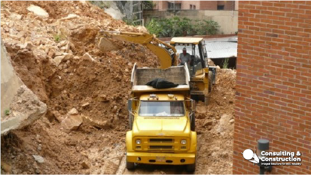 Una excavadora amarilla carga tierra en un camión volquete amarillo en una obra, donde los trabajadores se centran en la estabilización de ladera cerca de un edificio de ladrillo. Montones de tierra y la zona de Juanambú son el escenario de este proyecto en curso.