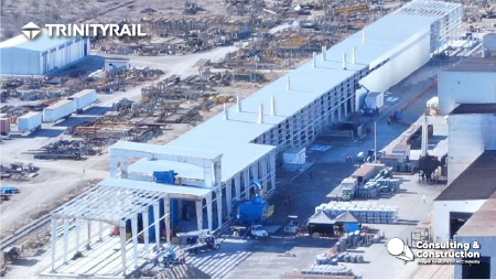 Vista aérea de una gran obra de construcción industrial en la que se está construyendo la Nave Trinity, un largo edificio blanco con estructura de acero. Maquinaria, materiales y vehículos están dispersos alrededor, con "TRINITYRAIL" y logotipos de la empresa superpuestos.