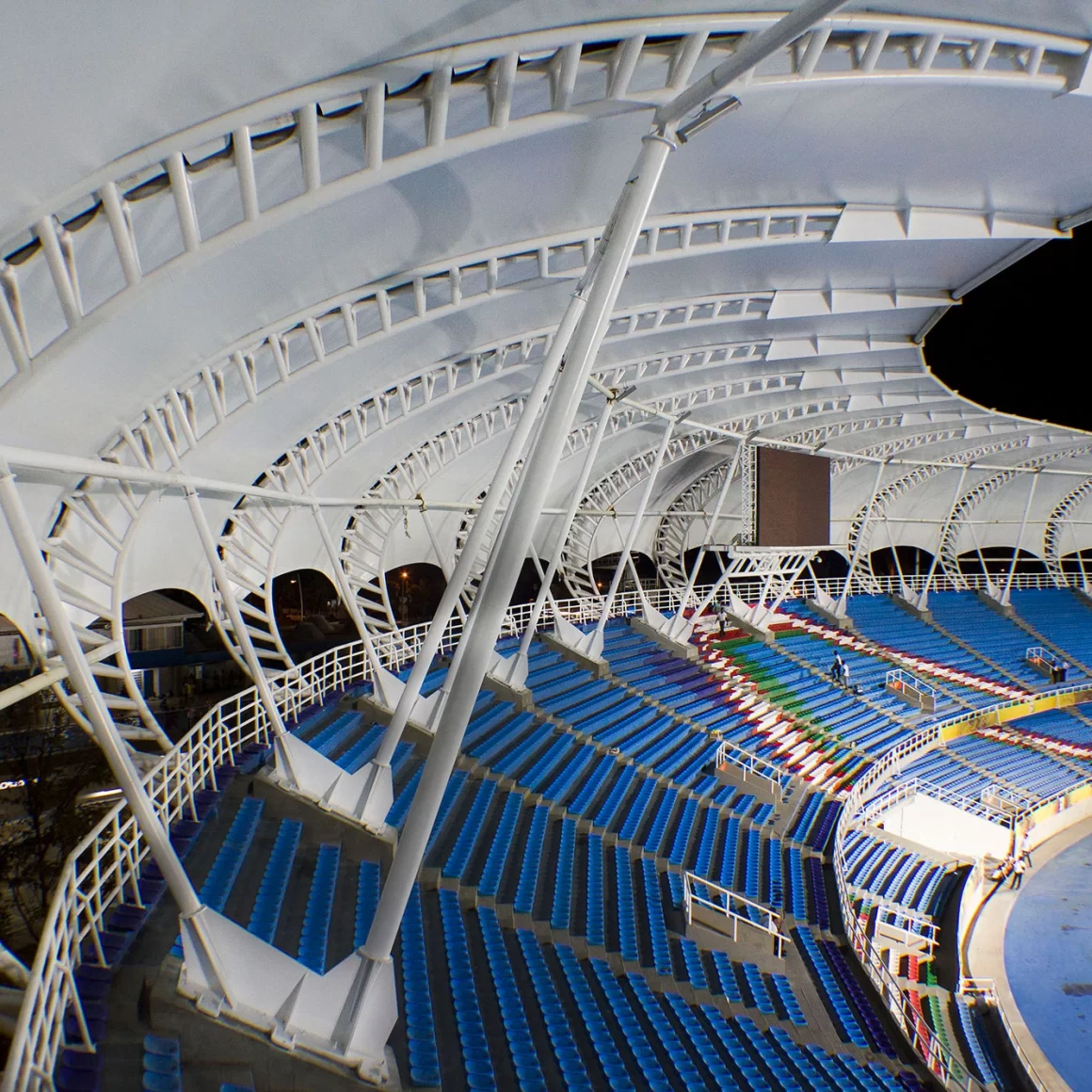 Un estadio grande y moderno con asientos azules y una característica estructura de cubierta blanca y curvada realizada con tensoestructuras para cubiertas arquitectónicas, fotografiado de noche. Las gradas están vacías y el campo es parcialmente visible.