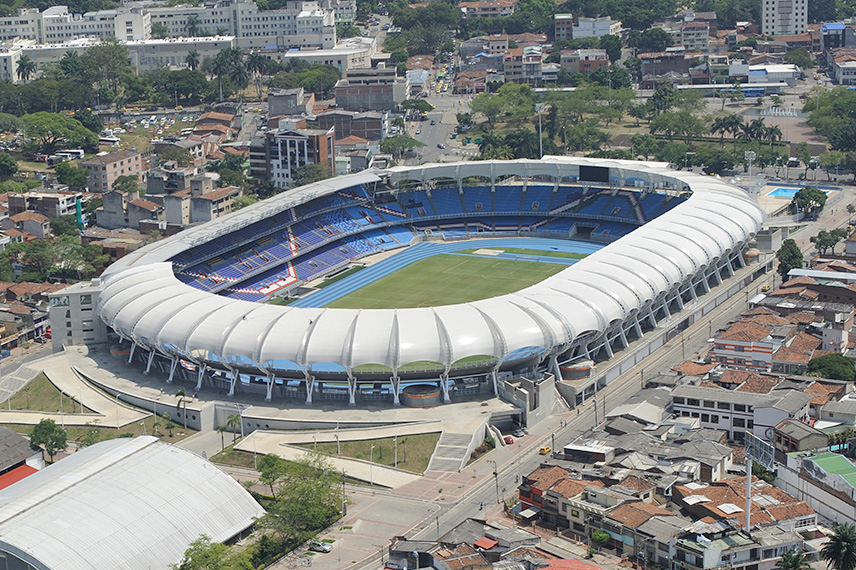 Fotografía aérea con drones para construcción Vista aérea de un gran estadio moderno con techo blanco y asientos azules, que rodea un campo de fútbol verde con una pista de atletismo. El estadio está situado en una zona urbana con edificios y árboles en las inmediaciones.